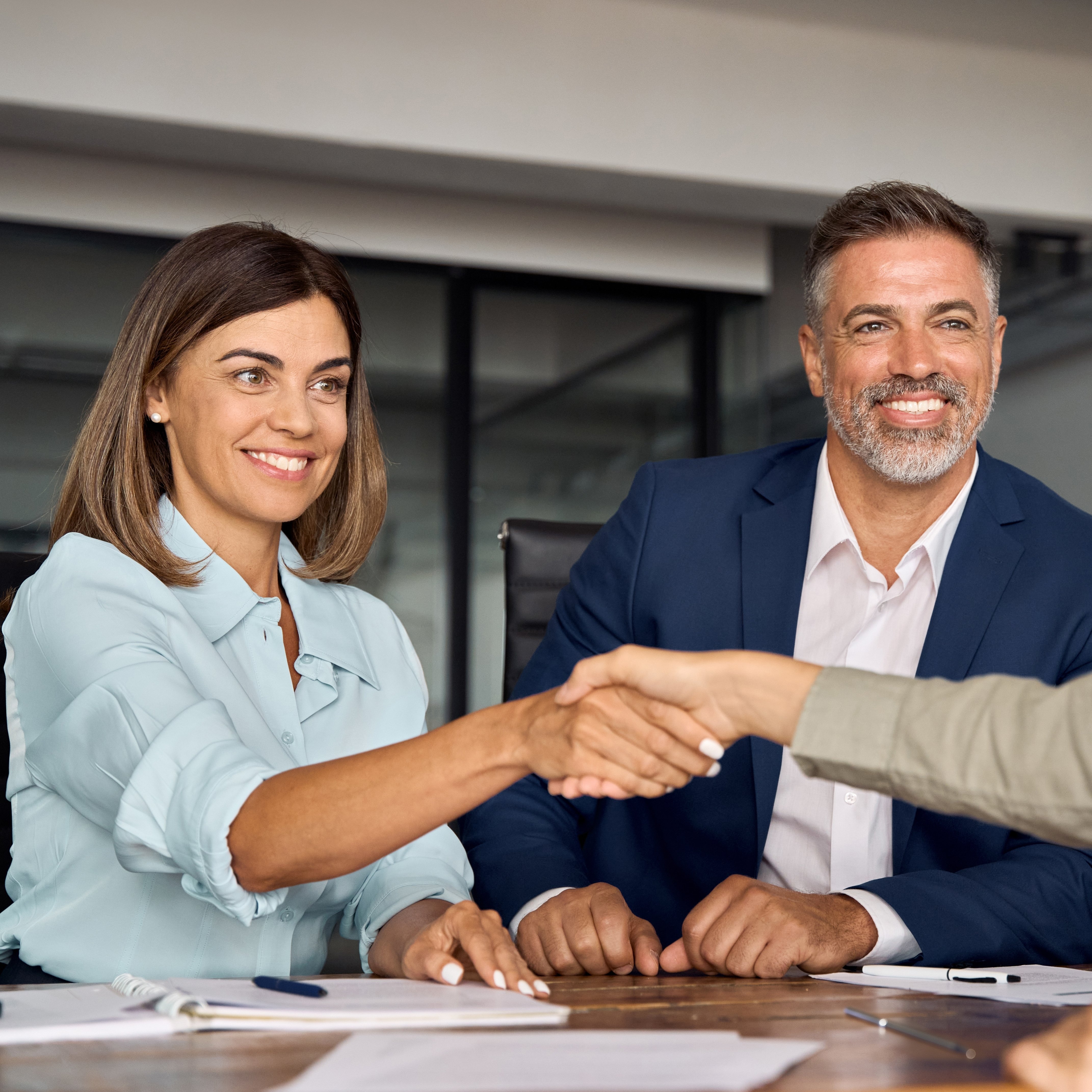 Situation Vertragsunterschrift: Mann und Frau auf einer Seite des Tisches. Eine Mitarbeiterin auf der anderen. Die beiden Frauen schütteln sich die Hand. Alle lächeln.