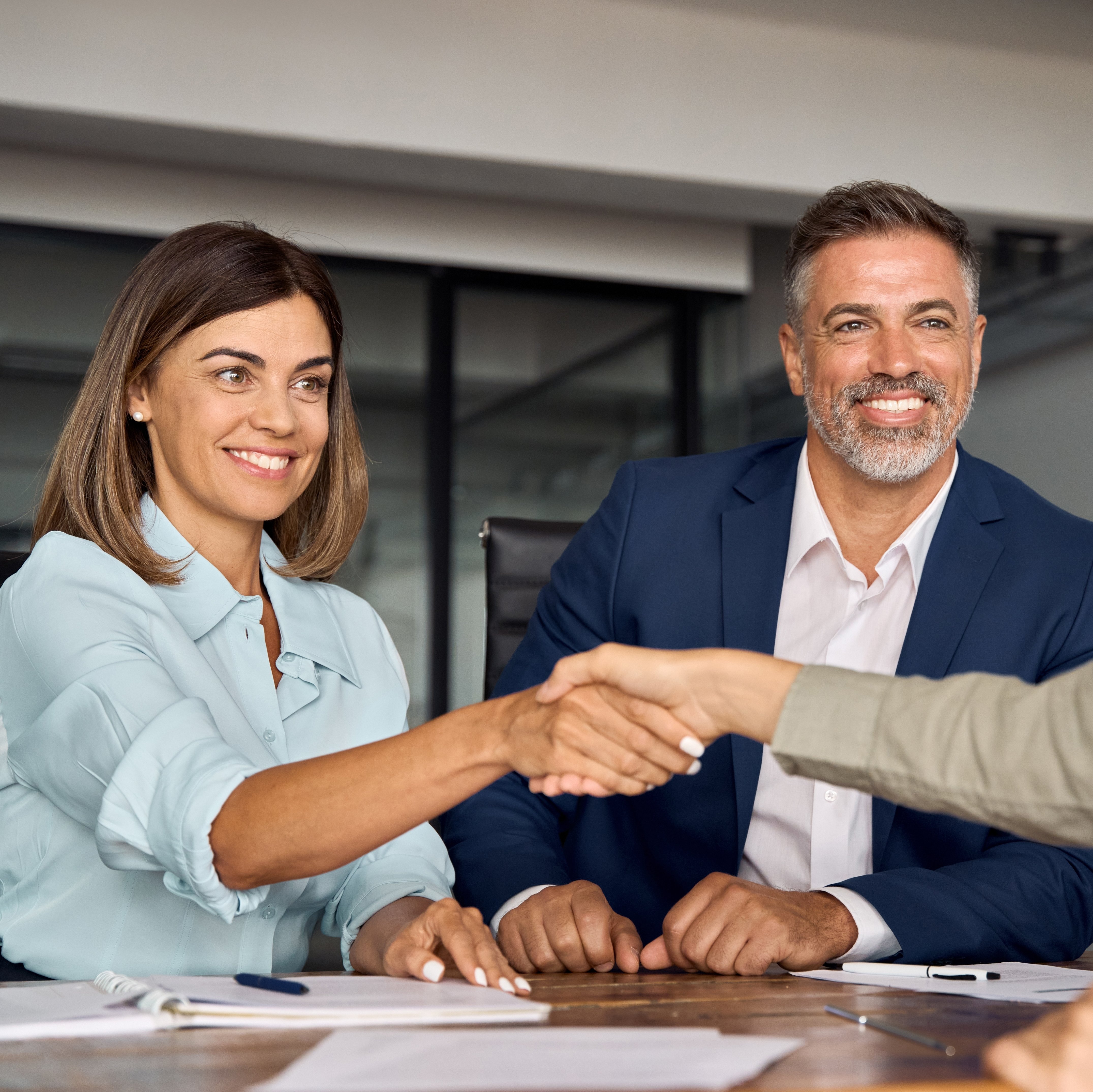 Situation Vertragsunterschrift: Mann und Frau auf einer Seite des Tisches. Eine Mitarbeiterin auf der anderen. Die beiden Frauen schütteln sich die Hand. Alle lächeln.